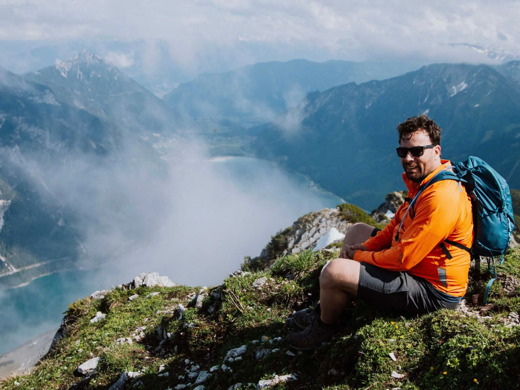 Gerd-Jan in een oranje jas met rugzak, zittend op een bergtop boven een meer tussen de wolken.